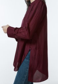 Burgunderfarvet letvægts skjorte med lange ærmer og afslappet pasform, der har en knappet sideslids og tekstureret stof. Båret sammen med blå jeans.