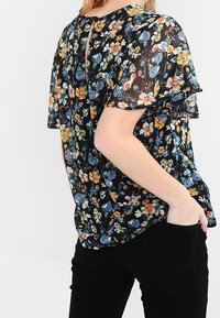 Žena v černé květované blůze s krátkými rukávy s volánky, v kombinaci s černými kalhotami, s rukou v kapse, otočená bokem.