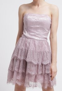 Ärmlös lila klänning med ett lager av spetskjol och slät satinklänning, bärs av en person med bara axlar och synliga armar.