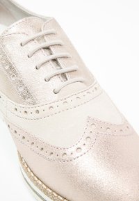 Gros plan d'une chaussure brogue rose clair brillante avec des détails perforés et des lacets assortis sur un fond blanc.