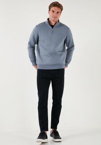 Felpa zip-up azzurro chiaro con polsini e fondo a costine, abbinata a jeans scuri e sneakers nere, in piedi contro uno sfondo neutro.