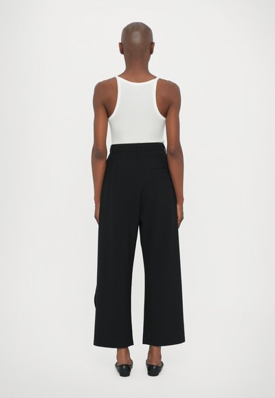 Black wide-leg trousers with a tailored fit, featuring a high waist, smooth texture, and a back pocket detail. Paired with a white tank top.