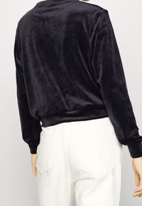 Svart sammets skjorta med långa ärmar och avslappnad passform, rundad bakkant och vida ärmar, som matchas med höga vita jeans med bakfickor.