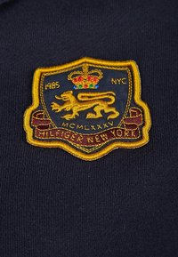 Marinblått tyg med broderad sköldlogotyp som föreställer ett lejon, en krona och texten "1985 NYC MCMLXXXIV HILFIGER NEW YORK" i guld och vinröd.