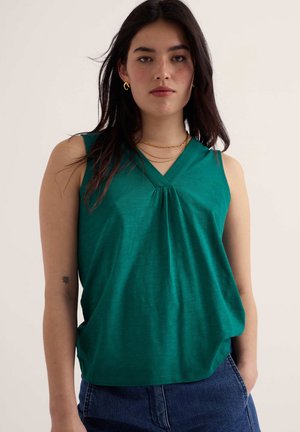 Žena s dlhými tmavými vlasmi, ktorá nosí zlaté kruhové náušnice, vrstvené zlaté náhrdelníky, tyrkysový bezrukávový top a modré džínsy, stojaca pred svetlým pozadím.