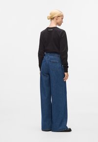 Sweatshirt noir à manches longues avec logo dans le dos, associé à un jean en denim bleu à jambe large avec deux poches arrière et des détails de couture visibles.