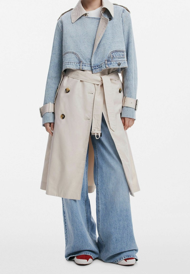 Jeansowy płaszcz w kolorze beżowym z krótką kurtką dżinsową na wierzchu, szerokie niebieskie dżinsy i sneakersy z czerwonymi akcentami.