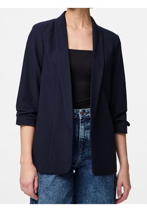 Kvinde iført en mørkeblå blazer med rynkede trekvart-ærmer over en sort top og højtaljede blå denimjeans mod en hvid baggrund.