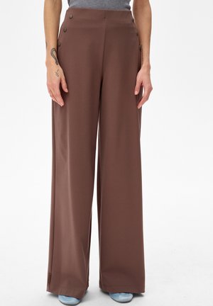 Kobieta ubrana w brązowe spodnie z wysokim stanem i szerokimi nogawkami, z guzikami po lewej stronie, zestawione z jasnoniebieskimi butami.