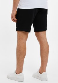Schwarze Sportshorts aus weichem Material, mit schlankem Schnitt und dezentem Logo-Detail am Saum. Kombiniert mit weißen Turnschuhen.
