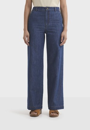 Personne portant un jean bleu taille haute à jambes larges, associé à des sandales ouvertes en daim beige, debout devant un fond clair uni.