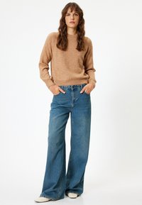 Maglione beige a maglia con scollatura a girocollo e polsini a coste, abbinato a jeans blu a vita alta e larghi e scarpe bianche.