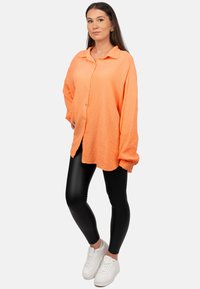 Camicia arancione oversize e strutturata con colletto, abbinata a leggins neri e sneakers bianche.