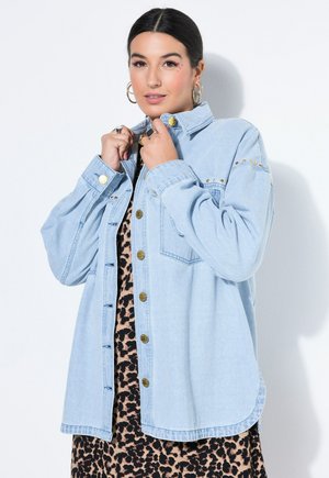 Kvinde retter på kraven af en lyseblå denimjakke, båret over en leopardmønstret kjole, med guldring-øreringe og ringe, foran en hvid baggrund.