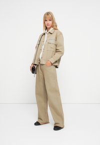 Beige denimjakke med fire lommer, paret med matchende vide bukser og svarte sko. Hvit skjorte under, minimalistisk design.