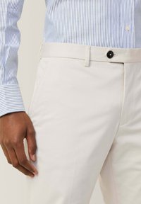 Crème plooibroek met een gladde textuur, voorzien van een enkele knoopsluiting en zijzakken, gecombineerd met een blauw-wit gestreept overhemdd.