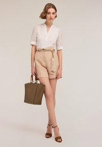 Witte, kortekogelige blouse met oogjesdetails, tan hoge taille shorts en een bruine tote bag met metallic accenten. Beige haksandalen.
