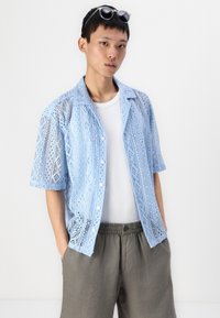 Lyseblå bluse i blonder med kort erm og geometriske mønstre, knappelukking, båret over en hvit t-skjorte og sammen med grå shorts.