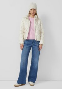 Kremowa pikowana kurtka z wysokim kołnierzem, różowy sweter i rozszerzane niebieskie dżinsy. Model nosi lekki dzianinowy kapelusz i casualowe buty z grubą podeszwą.