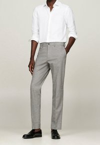 Pantaloni grigi su misura con una leggera trama e un motivo sottile, abbinati a una camicia bianca con bottoni, completati da scarpe nere.