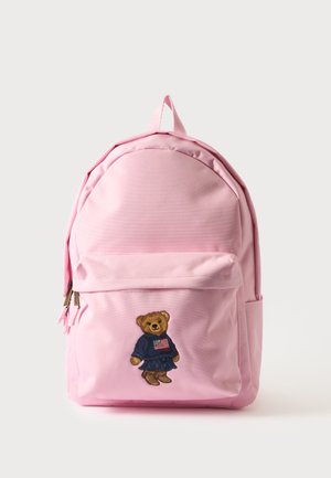 Sac à dos rose avec une poche avant brodée d'un ours en peluche portant un pull bleu marine et des bottes sur un fond clair uni.