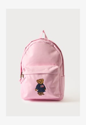 Sac à dos rose avec une poche avant brodée d'un ours en peluche portant un pull bleu marine et des bottes sur un fond clair uni.