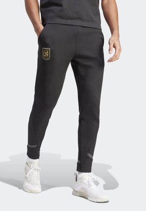 Man wearing black Los Angeles FC athletic pants and white sneakers, standing with hands in pockets against a plain white background.