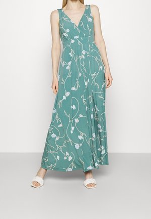 Vrouw draag een mouwloze teal maxi-jurk met witte bloemenpatronen en witte gevlochten open teen sandalen, staand tegen een witte achtergrond.