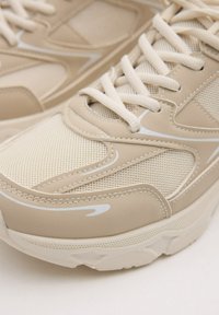 Las zapatillas deportivas beige cuentan con un upper de malla y sintético, un acabado texturizado, detalles en blanco y cordones planos, diseñadas para brindar comodidad y durabilidad.