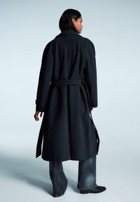 Tamna, duga kaput sa pojasom u struku, širokim rukavima i ovratnikom. Napravljen od teksturiranog materijala, uparen s širom trapericama i čizmama.