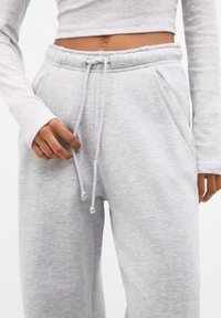 Helle graue Sweatpants aus weichem Stoff, mit einem Kordelzugbund, seitlichen Taschen und einem lockeren Schnitt. Die Oberfläche wirkt glatt.