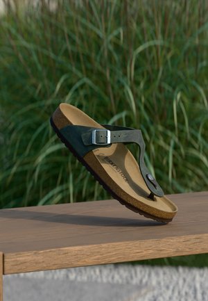 Fekete Birkenstock szandál csattos pánttal, amely egy fa felület fölött lebeg, háttérben zöld növényekkel.