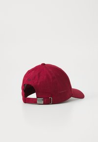 Red cap with a textured fabric, curved brim, adjustable strap at the back, and a small button on top. Features ventilation holes along the sides.