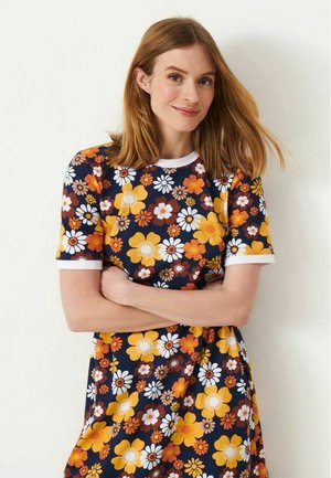 Kvinde med skulderlangt rødt hår iført en mørkeblå kjole med orange, gule og hvide blomster-mønstre, står med armene over kors.