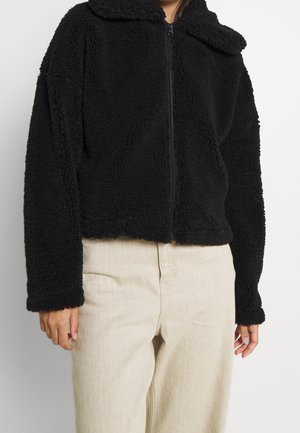 Persona con una chaqueta polar negra con cremallera y mangas largas y pantalones de tiro alto de color beige claro sobre un fondo liso.