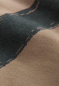 Strickstoff mit einer strukturierten Oberfläche und einem auffälligen dunkelgrünen abstrakten Design auf beigem Hintergrund, der weiches, warmes Material präsentiert.