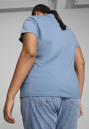 Person wearing a light blue short-sleeve t-shirt and blue jeans, standing with back facing the camera against a plain background.