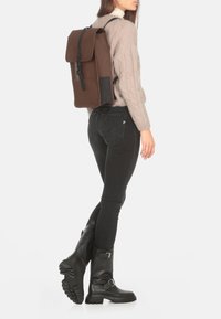 Mochila rectangular marrón con una solapa, correas ajustables y acentos negros. Llevada en la espalda, combinada con un suéter de punto y jeans negros.