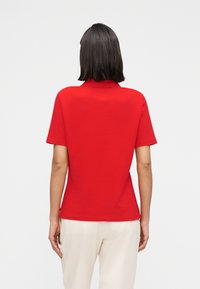  Γυναίκα με ίσια σκούρα μαλλιά, ντυμένη με απλό κόκκινο πόλο με κοντά μανίκια και άσπρο παντελόνι, εμφανίζεται από πίσω σε λευκό φόντο.