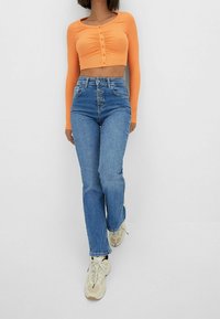 Oranžový dlouhý rukáv crop top s detaily knoflíků, v kombinaci s modrými džíny s vysokým pasem a béžovými teniskami. Jednoduchý, přiléhavý design.
