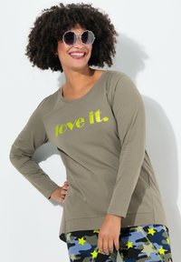 Oliivinvihreä pitkähihaisen paidan, jossa lukee "rakasta sitä." kirkkaan keltaisella, yhdistettynä kuvioituihin sinisiin maastohousuihin, joissa on keltaisia tähtiä.