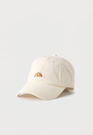 Off-white baseballkasket med en lille broderet brun croissant foran i midten, vist mod en ensfarvet baggrund.