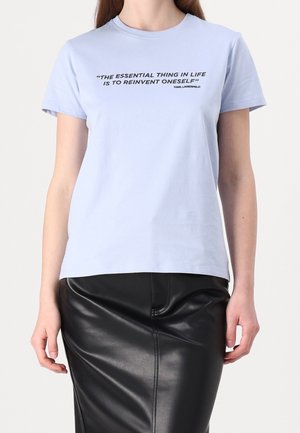 Vrouw die een lichtblauw T-shirt draagt met de tekst "Het essentiële in het leven is jezelf steeds opnieuw uitvinden" en een zwarte leren rok.