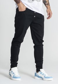 Vaqueros de mezclilla negros de corte slim, con cinco bolsillos y cierre de botón. Combinados con zapatillas altas en blanco con acentos en azul.