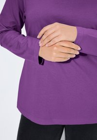 Lila Langarmshirt mit glatter Textur und Rundhalsausschnitt. Die Ärmel haben ein leichtes Aufschlagdetail. Getragen mit dunklen Jeans.