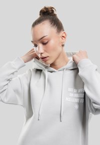 Sudadera gris claro hecha de tela suave, que cuenta con un bolsillo frontal, cordones y texto bordado con un gráfico de sol en el pecho.