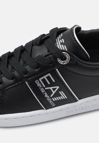 Black synthetic leather sneaker with white rubber sole, flat laces, and logo accents. Features a smooth surface and stitched detailing.