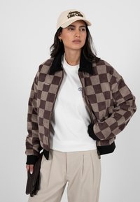 Femme portant une casquette beige, une veste à carreaux marron, un t-shirt blanc, un pantalon beige, tenant des gants marron foncé, debout devant un fond uni.