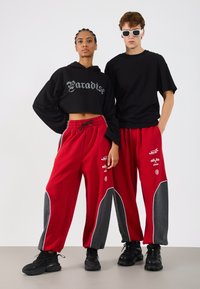 Crna kratka hoodie majica s printom "Paradise", crvene trenirke s sivim bočnim panelima i grafičkim akcentima, u kombinaciji s crnom obućom i sunčanim naočalama.