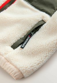 Rukáv krémovej fleecovej bundy s elastickým manžetovým lemom a zelenou zipsovou kapsou, ktorá má čiernu ťahaciu slučku Superdry.